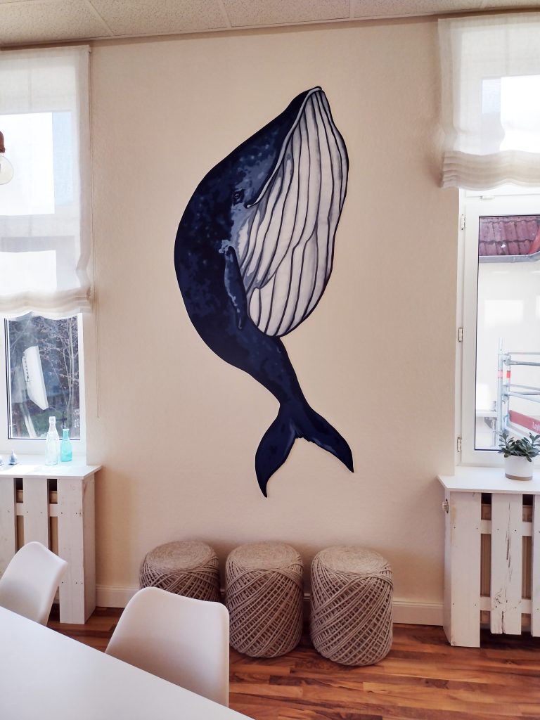 Ein großes Wal-Wandbild an einer beigen Wand über drei geflochtenen Hockern in einem hellen Raum.