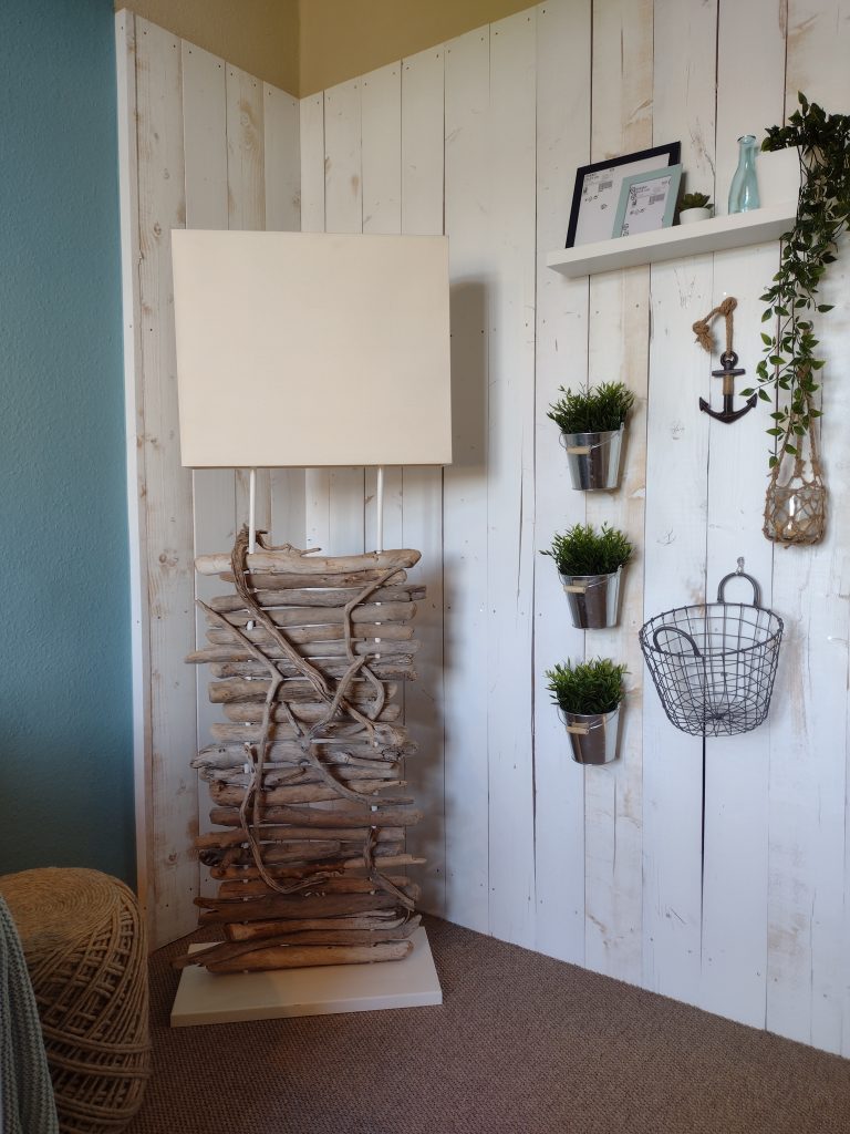 Stehlampe mit einem Sockel aus Treibholz neben einer weißen, holzgetäfelten Wand mit Dekorationsartikeln.