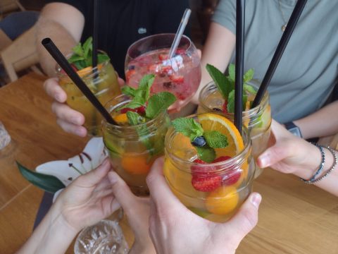 Fünf Personen stoßen mit Strohhalmen und Minzblättern auf einem Holztisch auf Gläser mit Fruchtcocktails an.