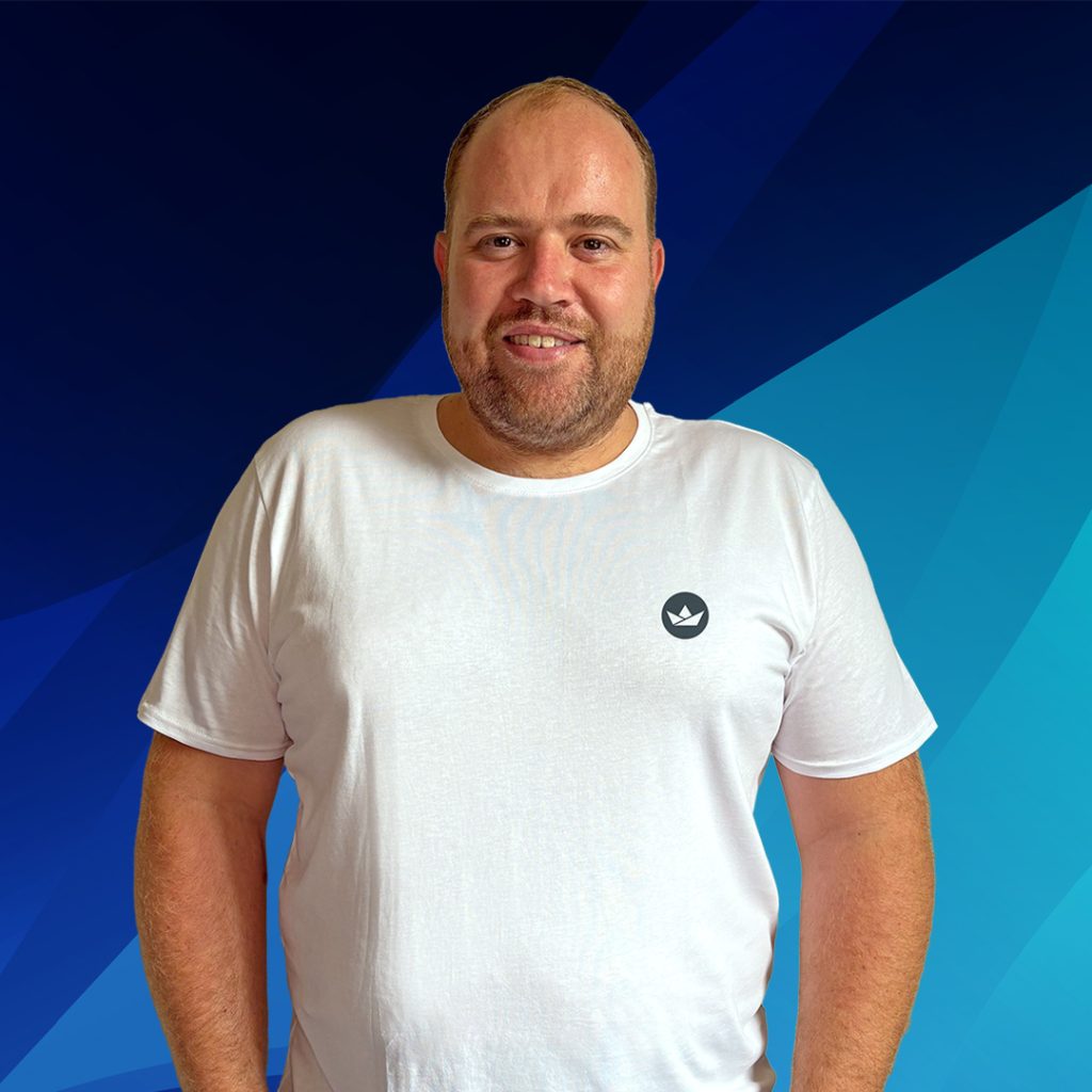 Ein Mann in einem weißen T-Shirt steht vor einem blauen, geometrischen Hintergrund und lächelt in die Kamera.