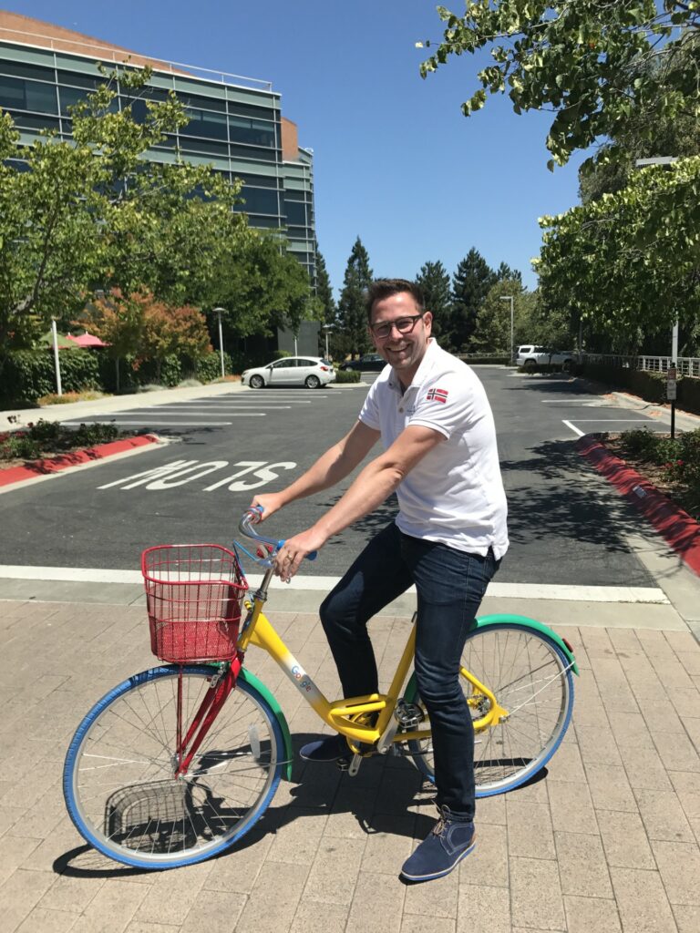 Mann im weißen Hemd auf einem bunten Fahrrad an einem sonnigen Tag in der Nähe von Bürogebäuden.