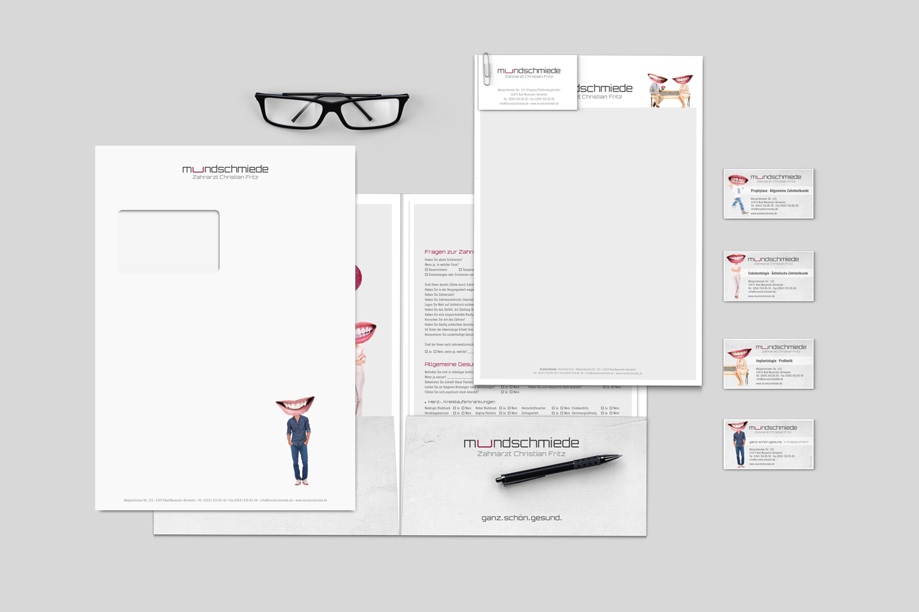 Briefpapier-Set mit Briefkopf, Umschlägen, Visitenkarten, Brille und einem Stift auf grauem Hintergrund.