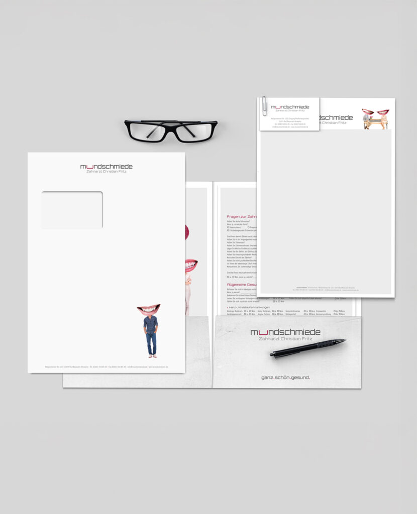 Büromaterialset mit Gläsern, Umschlägen, Visitenkarten und Briefpapier auf einer grauen Oberfläche.
