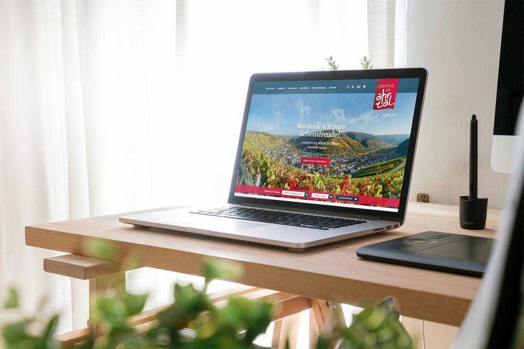 natuerlich-im-ahrtal_website Laptop auf einem Holztisch, auf dem eine Website mit einem Weinberg und Hügeln zu sehen ist.