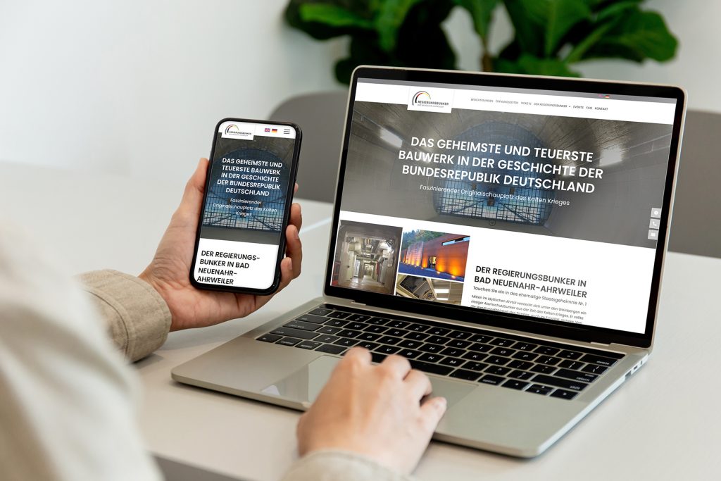 regierungsbunker_web-flat Person mit einem Smartphone und einem Laptop, die beide die gleiche deutsche Website anzeigen.