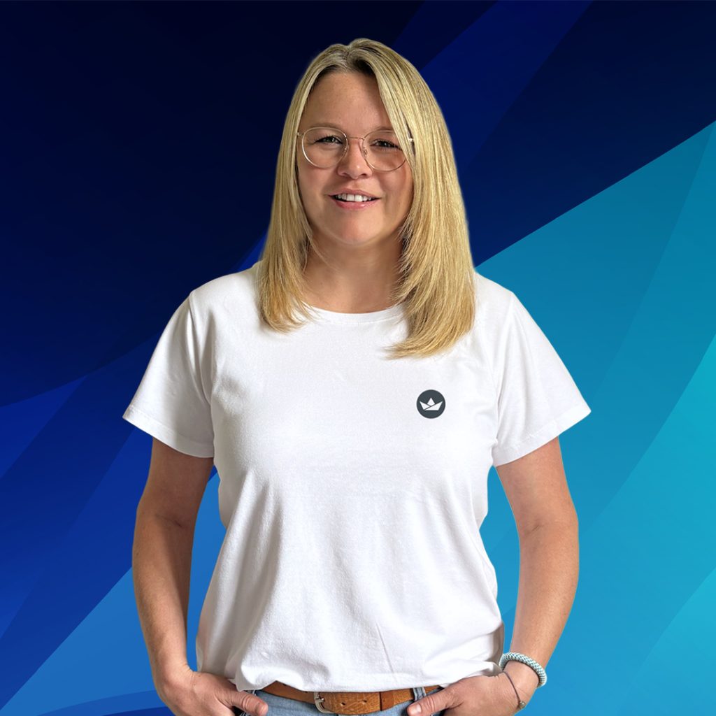 Eine Frau mit langen blonden Haaren, Brille und weißem T-Shirt steht vor einem blauen Hintergrund.