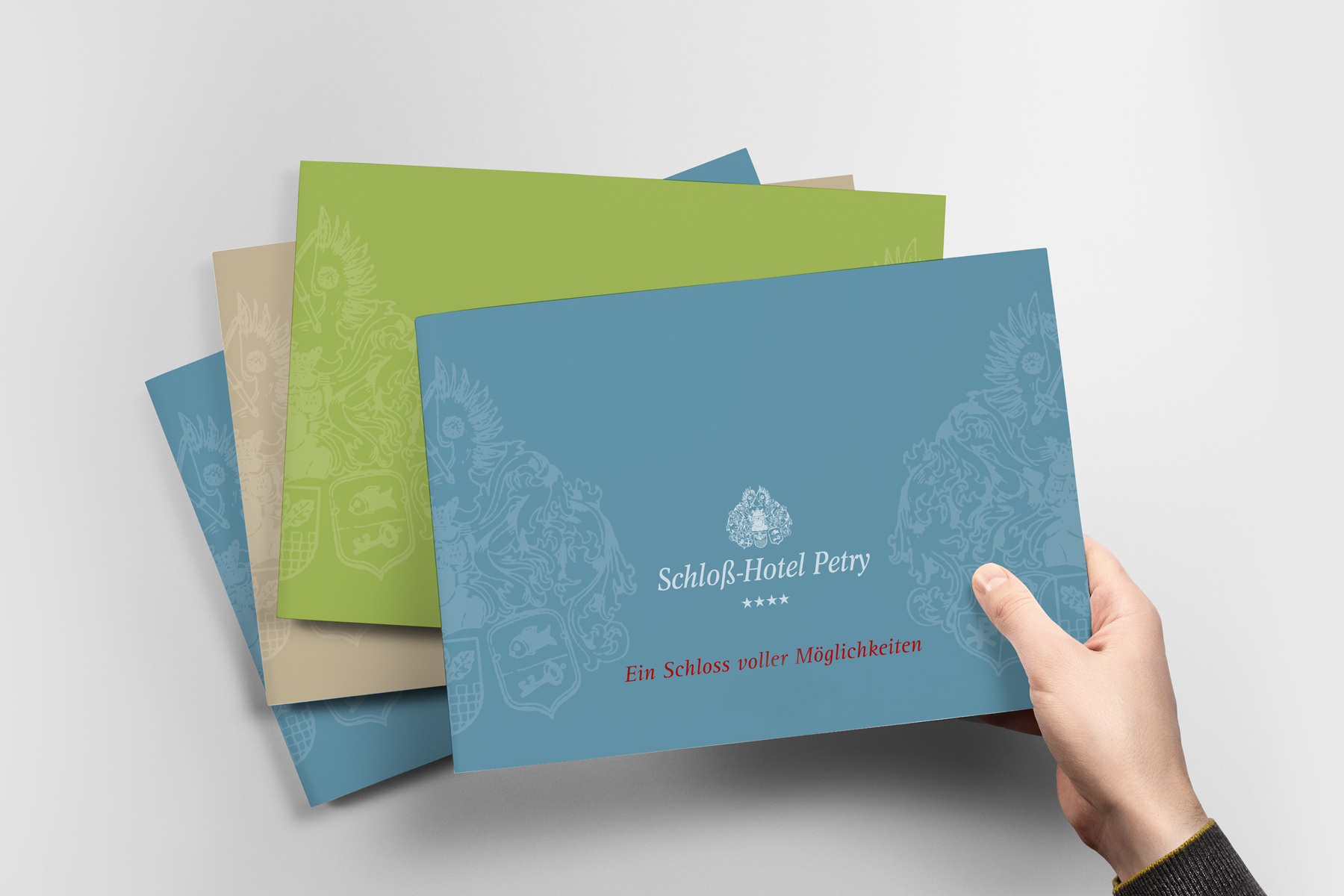 Eine Hand hält eine blaue Broschüre des Schloss-Hotels Petry, unter der sich weitere in Grün und Beige befinden.