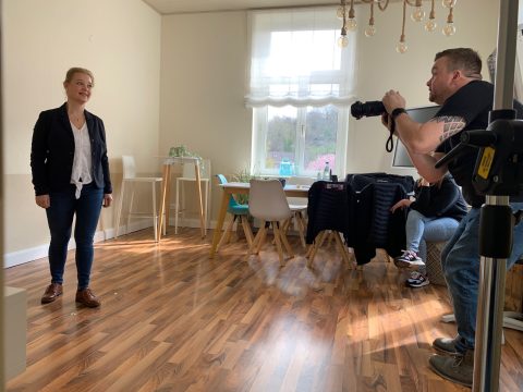 Ein Fotograf nimmt ein Porträt einer Frau auf, die in einem hell erleuchteten Raum mit Holzboden steht.