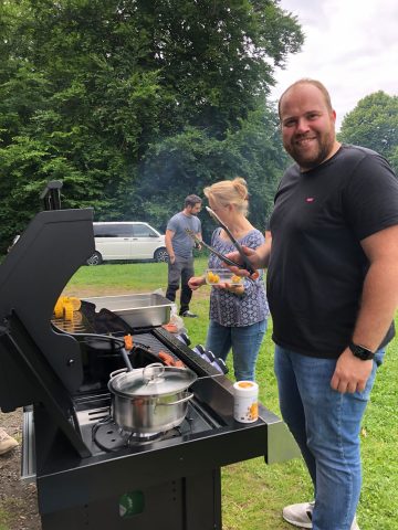Ein Mann grillt sein Essen im Freien, während andere in der Nähe auf einer Wiese stehen.