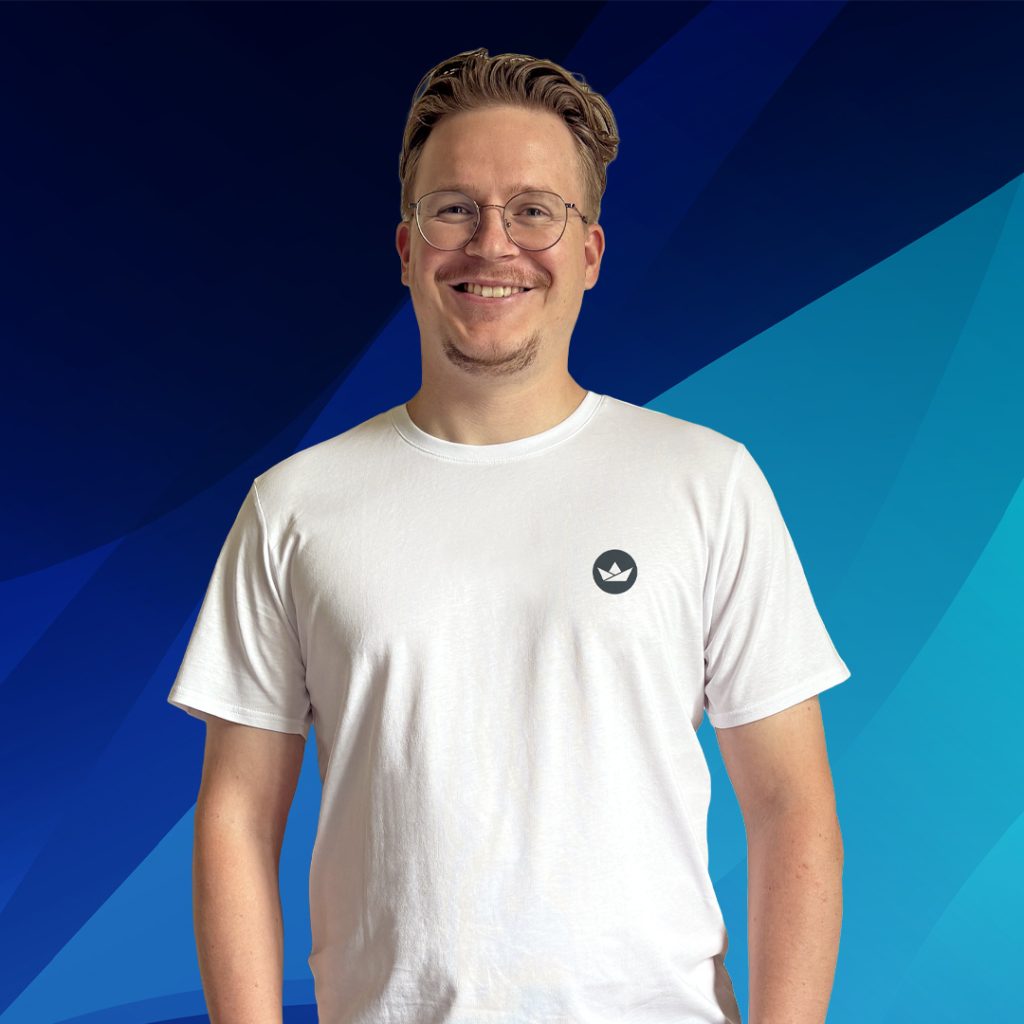 Ein lächelnder Mann mit Brille und weißem T-Shirt steht vor einem blauen, geometrischen Hintergrund.