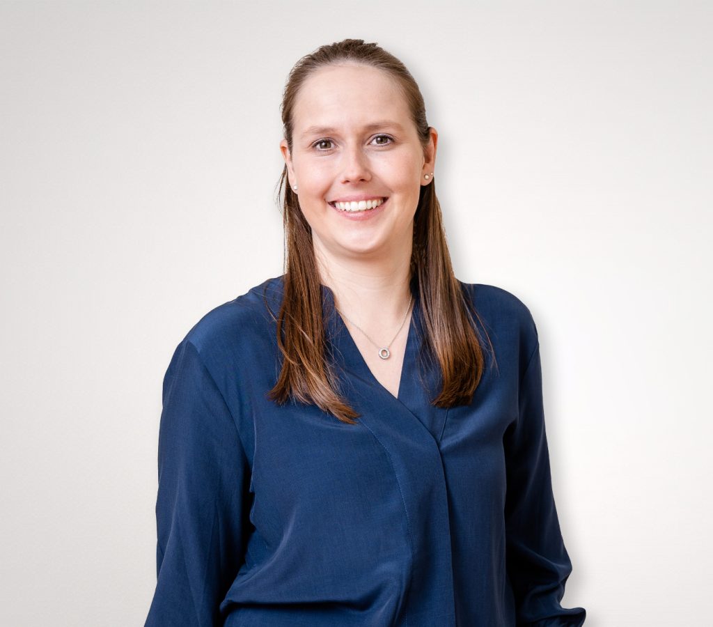 verena Frau mit langen braunen Haaren in einer blauen Bluse steht und lächelt vor einem schlichten Hintergrund.