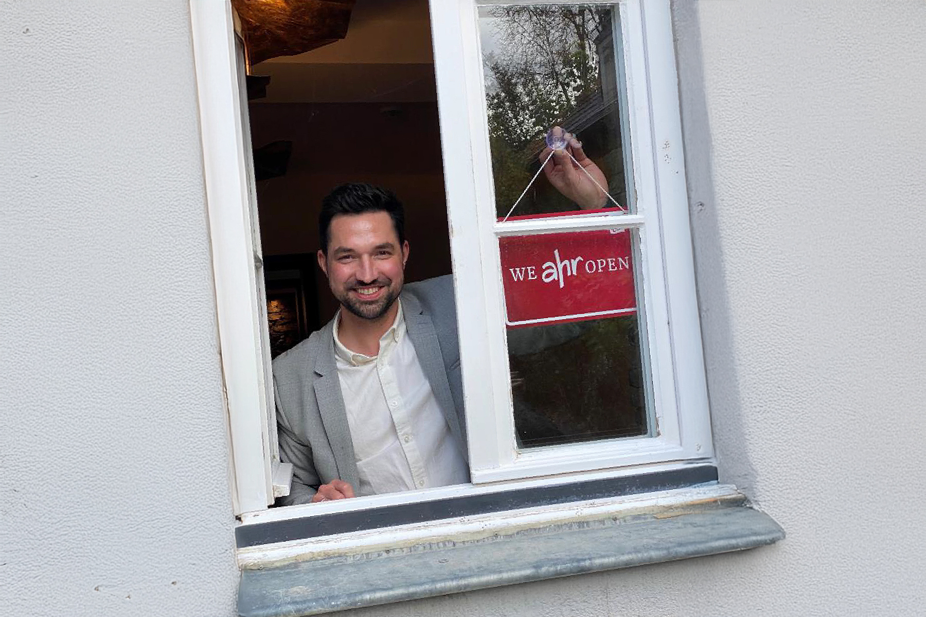 Ein Mann im Anzug öffnet ein Fenster und hält ein "Wir haben geöffnet"-Schild hoch.