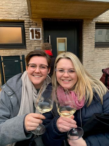 Zwei Frauen lächeln und halten Weingläser vor einem Haus mit der Nummer 15.