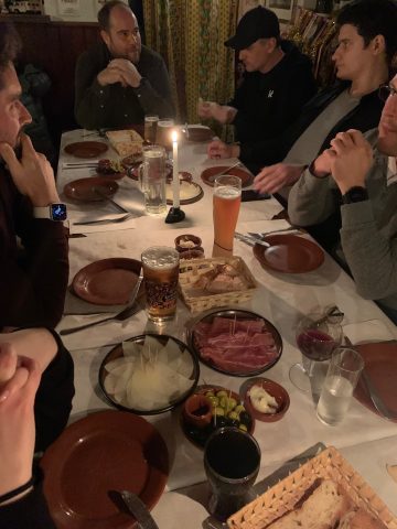 Sechs Personen sitzen um einen Tisch mit Getränken, Brot und Vorspeisen, der von einer einzigen Kerze beleuchtet wird.