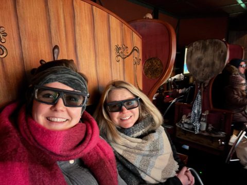 Zwei Frauen mit 3D-Brillen sitzen auf hölzernen Sitzen in einer Indoor-Attraktion.