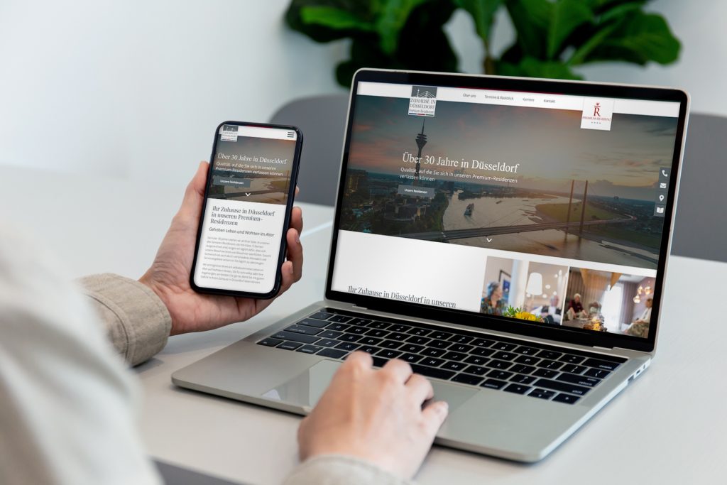 zuhause-in-duesseldorf_web-flat-L Eine Person, die dieselbe Unternehmenswebsite auf einem Smartphone und einem Laptop am Schreibtisch betrachtet.